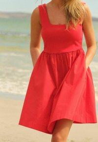 Robe sans manches rouge avec un décolleté carré, une jupe évasée et des poches. Tissu lisse, longueur genoux et légère pour un temps chaud.
