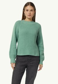 Hellgrüner, gerippter Strickpullover mit Rundhalsausschnitt, langen Raglanärmeln und strukturiertem Muster. Kombiniert mit dunklen Hosen.