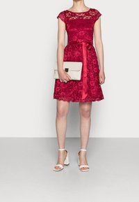 Röd spetklänning med ärmlösa cap sleeves och en vågad fåll, med ett sidenbälte. Matchas med en beige kuvertväska och vita sandaler med blockklack.