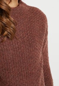 Detail osoby nosící texturovaný hnědý pletený svetr s žebrovaným vysokým límcem a částečně viditelnými dlouhými vlnitými vlasy.