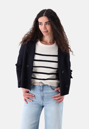 Woman with long curly hair wears navy double-breasted jacket, white sweater with black stripes, light blue jeans, and beige belt, hands in pockets.