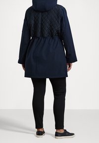Navy blue coat with a quilted black back, featuring a hood, drawstring waist, and long sleeves, paired with black pants and sneakers.