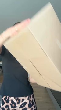 Brūna kartona kaste, kuru tur rokās. Kaste ir taisnstūra veida, tai nav redzamu uzlīmju vai atzīmju, un augšdaļa ir neizsieta.