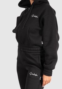 Black zip-up hoodie and matching joggers made of soft fabric, featuring elasticized cuffs and waistband, with a white logo on the chest.