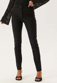 Femme portant un pantalon noir ajusté, un haut en dentelle noire à manches longues avec des poignets évasés, et des sandales à talons ouverts noires, se tenant devant un fond uni.