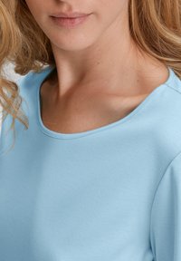 Camicetta azzurro chiaro a maniche corte con scollatura rotonda, realizzata in tessuto morbido. La texture sembra liscia con cuciture sottili sulle spalle.