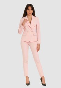 Donna che indossa una giacca doppiopetto rosa chiaro e pantaloni slim coordinati, in piedi con una mano vicino al colletto, tacchi neri.