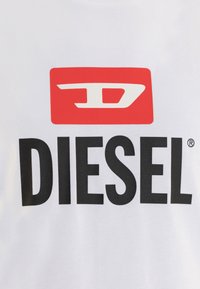 Balta T-kreklu ar lielu sarkanu taisnstūra logotipu, kurā ir stilizēts balts burts "D" un vārds "DIESEL" biezā melnā tekstā zem tā.