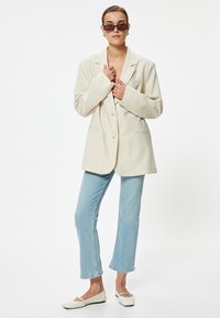 Blazer crema de corte oversize con dos botones, combinado con jeans rectos de color azul claro y bailarina blanca. Los accesorios incluyen unas gafas de sol negras y delgadas.