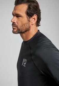 Černé sportovní tričko s kulatým výstřihem, dlouhými rukávy a mírně texturovaným povrchem. Na levé straně hrudi se nachází bílé logo.