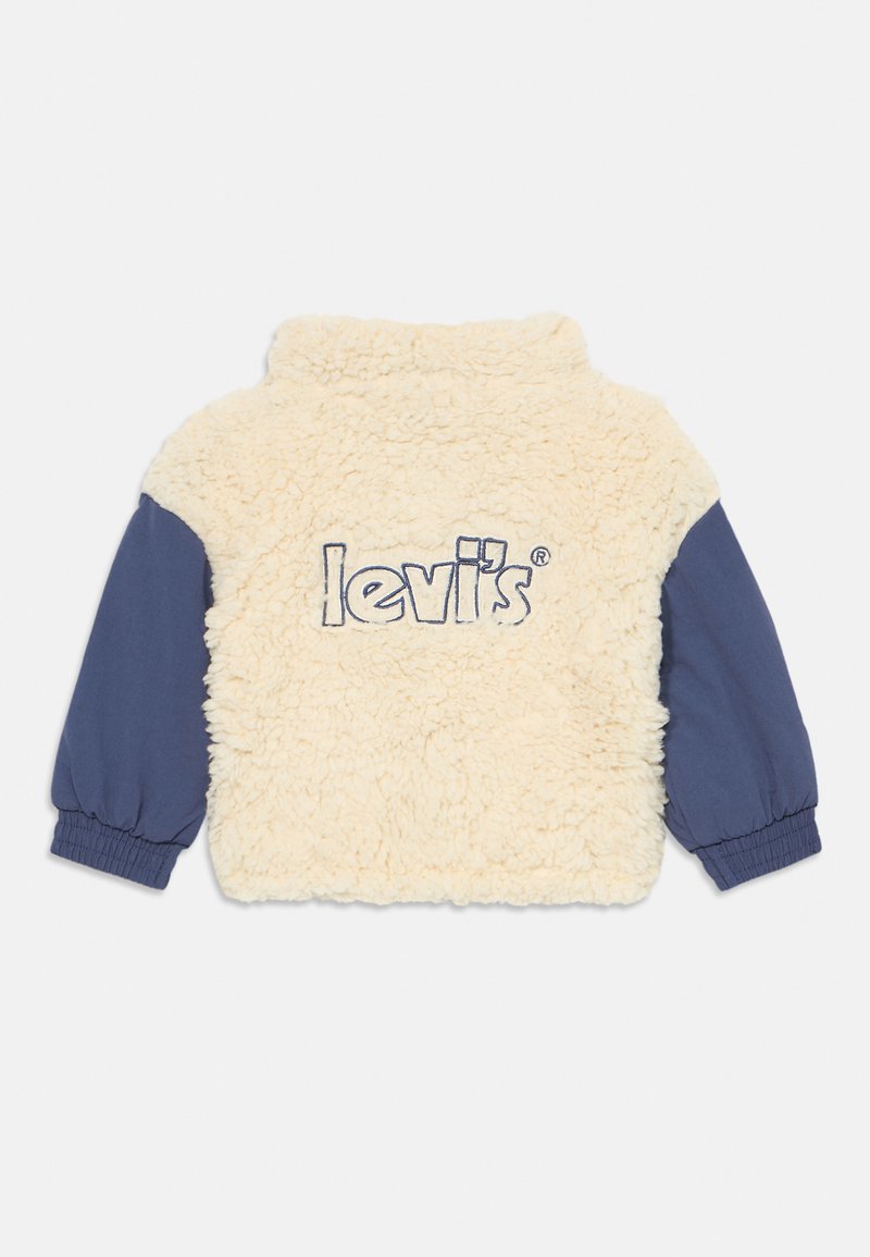 Chaqueta de forro polar tipo sherpa color crema con mangas largas azules y "Levi's" bordado en el centro de la parte posterior.