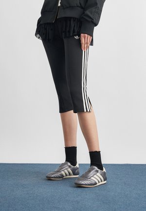 Osoba ubrana w czarne, krótkie spodnie Adidas z białymi paskami, czarne skarpetki, szare buty sportowe Adidas i czarną kurtkę, stojąca na niebieskim dywanie.