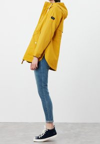Żółta wodoodporna kurtka z kapturem, przednią kieszenią i zapięciem na zatrzaski. Stylizowana z niebieskimi dżinsami i granatowymi trampkami na neutralnym tle.