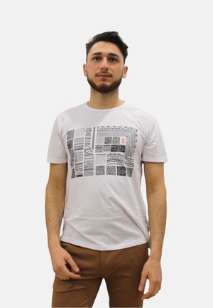 Giovane uomo con capelli corti e scuri e barba che indossa una maglietta bianca con quadrati neri decorati e pantaloni marroni, in piedi davanti a uno sfondo semplice.
