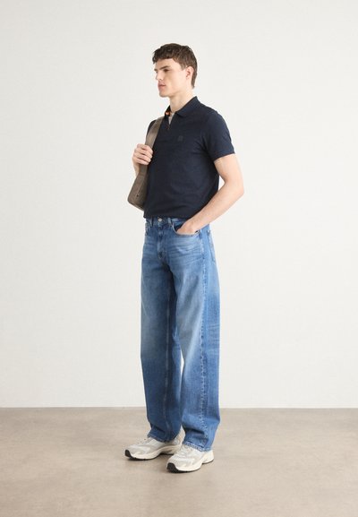 Polo shirt marine à manches courtes, jeans larges bleu clair, et baskets grises. Sac à bandoulière beige porté sur un bras.