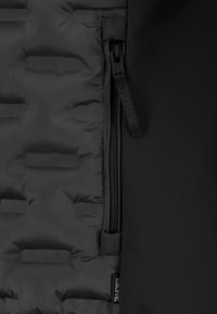 Schwarze gepolsterte Jackenstoff mit horizontaler Naht neben glattem Stoff, ausgestattet mit einem vertikalen Reißverschluss mit einem schwarzen Zugband, das mit "Sublevel" beschriftet ist.