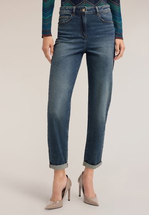 Frau trägt hochgekrempelte dunkelblaue Jeans und beige High Heels, mit einem gemusterten langärmligen Oberteil, das an der Taille sichtbar ist.