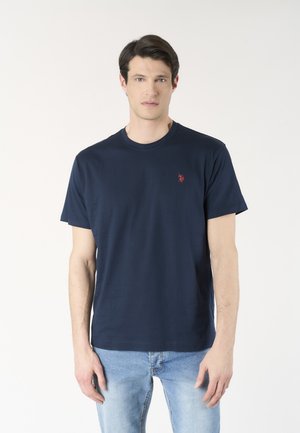 Uomo che indossa una maglietta blu navy semplice a maniche corte con un piccolo logo rosso sul petto e jeans azzurri, in piedi davanti a uno sfondo bianco.