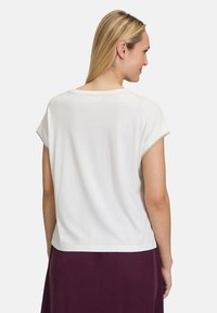 Haut blanc à manches courtes avec un col rond et des finitions en dentelle sur les manches, porté sur une jupe violette foncée. Texture lisse, coupe décontractée.
