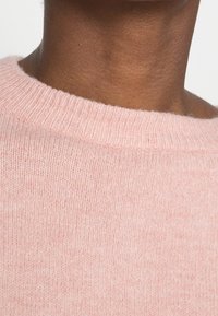 Pull rose clair en maille avec un col rond côtelé et un tissu doux et texturé, présentant une finition lisse et des variations subtiles de teinte.