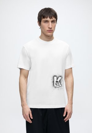 Jeune homme portant un t-shirt blanc à manches courtes avec une grande lettre noire "K" et un motif graphique en bas à droite.