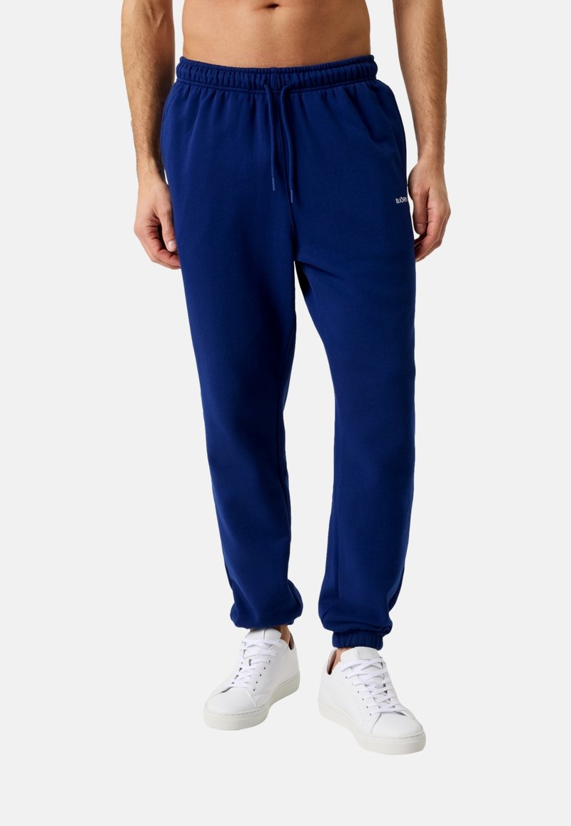 Muž oblečený v tmavomodrých teplákoch so šnúrkou a bielych teniskách, stojaci pred jednoduchým svetlým pozadím.