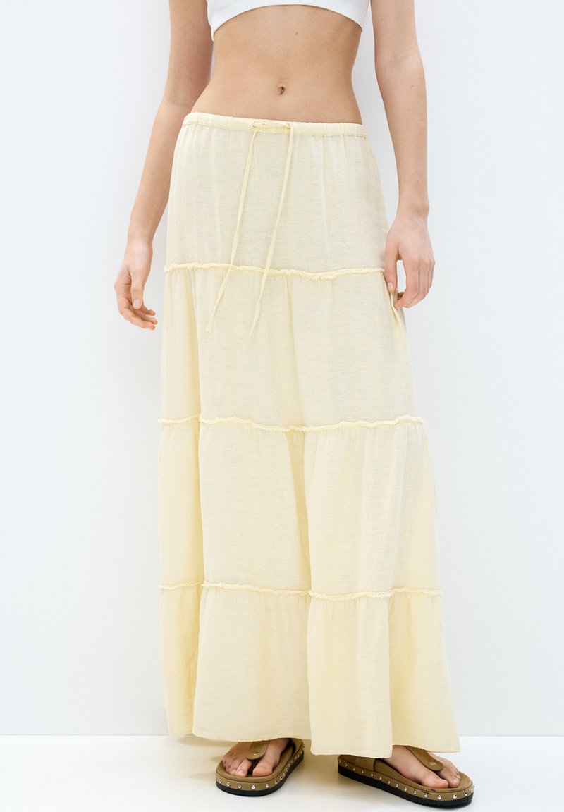 Jupe maxi tiered jaune clair en tissu doux, avec une taille à cordon de serrage et des détails froncés à chaque niveau. Portée avec des sandales plates.