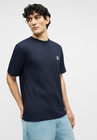 Νέος άνδρας με σγουρά μαλλιά που φοράει μπλε ναυτικό μπλουζάκι με μικρό λευκό λογότυπο στο στήθος και ανοιχτό μπλε τζιν, στέκεται μπροστά σε απλό φόντο.