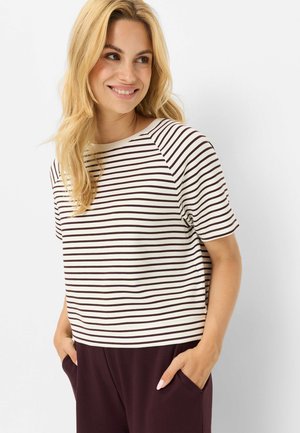 Blonde vrouw die een wit en donker gestreept T-shirt met korte mouwen en een donkere broek draagt, met handen in de zakken, glimlachend en naar rechts kijkend.