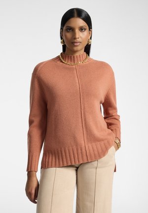 Kvinde med glat sort hår iført gyldne hoop-øreringe, et guldkæde-halskæde, en orange ribbet sweater og beige bukser med den ene hånd i lommen.