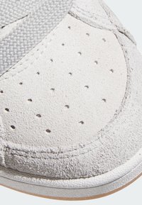 Hvite semskede sneakers med grå stoffstropp, sirkulære perforeringer, teksturert tå og gummisåle. Synlige sømmer langs kanten.