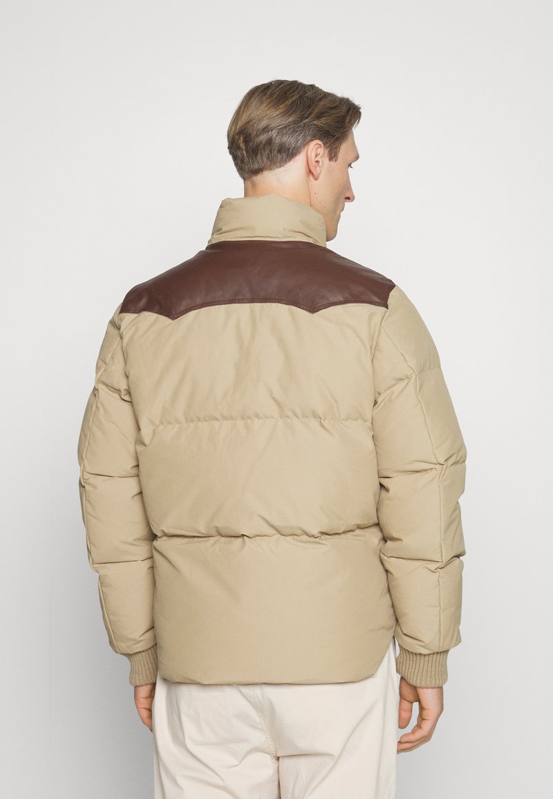Giacca puffer beige con spalle in pelle marrone, polsini a coste e colletto alto. Cuciture visibili e design imbottito.