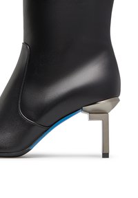 Stivaletto alla caviglia in pelle nera con un design elegante, caratterizzato da un tacco geometrico argentato e un accento blu sulla suola. Texture liscia.