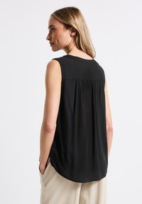 Černý bezrukávový top s kulatým výstřihem, splývavým materiálem a jemným skládáním na zadní straně. Ve spojení se světle béžovými kalhotami, které mají uvolněný design.