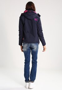 Marineblå vandtæt jakke med hætte i pink accent og logo, kombineret med blå jeans og hvide sneakers. Glat tekstur og funktionelt design.