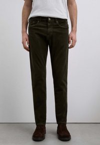 Pantalones de pana verde oscuro con un corte ajustado, que cuentan con cinco bolsillos y un cierre frontal de botón, combinados con botas de ante marrón.