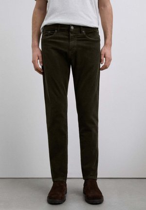 Pantalones de pana verde oscuro con un corte ajustado, que cuentan con cinco bolsillos y un cierre frontal de botón, combinados con botas de ante marrón.