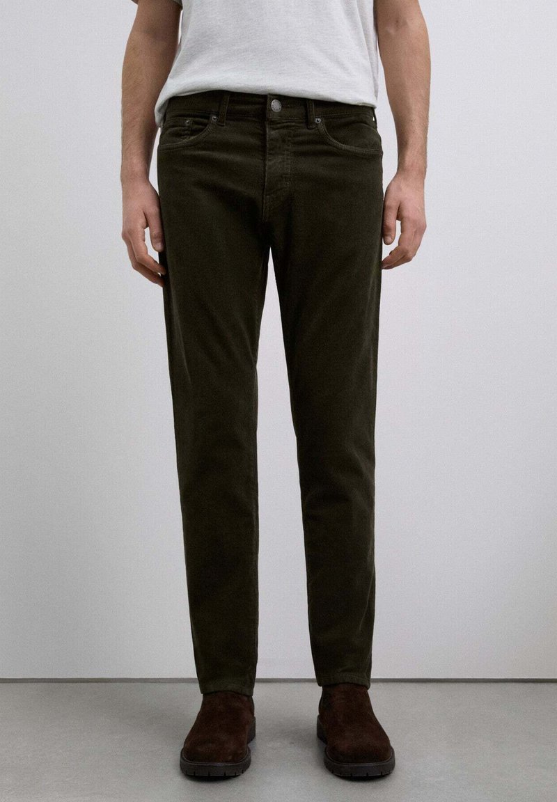 Pantalones de pana verde oscuro con un corte ajustado, que cuentan con cinco bolsillos y un cierre frontal de botón, combinados con botas de ante marrón.