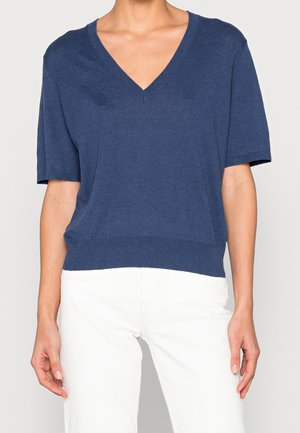 Osoba ubrana w granatowy sweter z krótkim rękawem i dekoltem w serek oraz białe spodnie, stojąca na tle jednolitego jasnego tła.