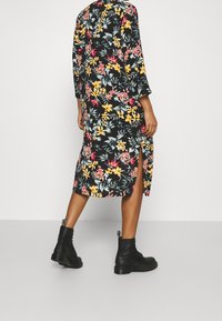 Blumenkleid in Midi-Länge mit schwarzem Hintergrund, das gelbe, rosa und grüne Muster aufweist. Hohe Schlitze an beiden Seiten. Ein Paar schwarze Knöchel-Stiefel.