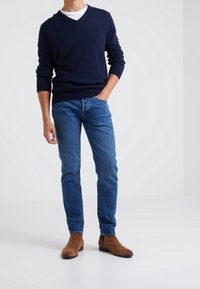 Maglione navy a scollo a v, camicia leggera sotto, jeans blu e stivali alla caviglia in suede marrone. Fit slim, polsini a costine e textures morbide.