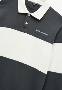 Poloshirt in Dunkelgrau und Weiß mit horizontalen Streifen. Mit einem weißen Kragen, drei Knöpfen und gesticktem Text "Einfach ist zeitlos."