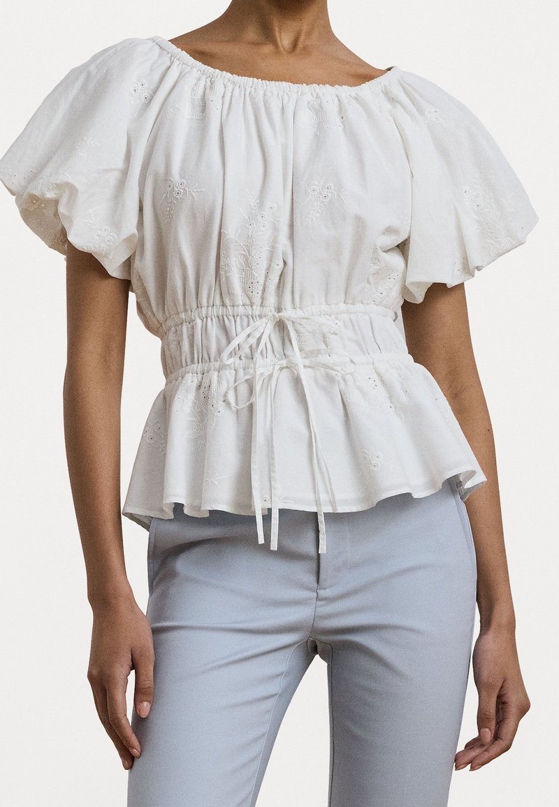 Vit, broderad blus med puffärmar, åtstramad midja och peplumslut. Har en rynkad halsringning och snörningsdetalj.