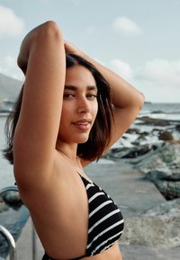 Top bikini a righe bianche e nere con un design triangolare; la modella ha i capelli di lunghezza media; lo sfondo presenta una scogliera rocciosa e il cielo.