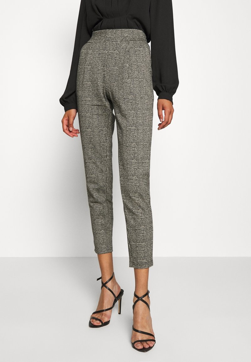 Femme portant un pantalon gris à carreaux taille haute et fuselé, un chemisier noir à manches longues et des sandales noires à talons hauts et à brides, debout sur un sol blanc.
