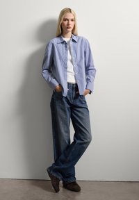Camisa de botón a rayas azul y blanca, material de algodón, con un bolsillo en el pecho, combinada con unos vaqueros de denim oscuro de corte holgado y zapatos marrones.
