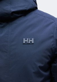 Veste bleu foncé avec capuche, logo HH embossé au dos près de l'épaule, détails de couture visibles sur le tissu.