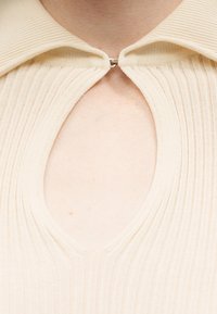 Εκρού ριμπ πλεκτό τοπ με γιακά που κλείνει με κλιπ και άνοιγμα σε σχήμα κλειδαρότρυπας στο στήθος, αποκαλύπτοντας γυμνό δέρμα από κάτω.