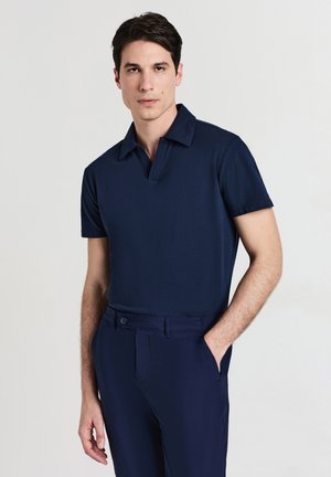 Homme portant une chemise polo à manches courtes bleu marine et un pantalon bleu marine assorti, se tenant avec une main dans la poche sur un fond neutre.