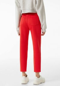 Jeans de mezclilla rojos con una cintura ajustada, piernas rectas, dos bolsillos traseros y un corte recortado, combinados con zapatillas blancas.